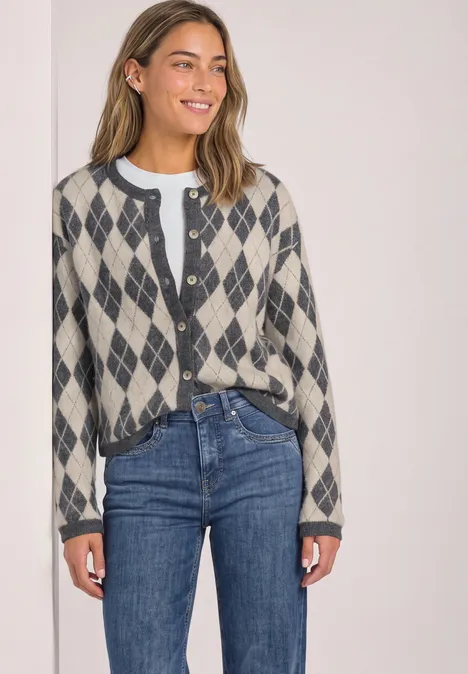 Eine Frau lächelt und trägt einen graubeigen Cardigan mit Rautenmuster, darunter ein weißes Shirt und blaue Jeans.