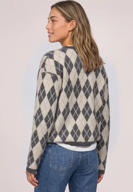 Frau von der Seite in grauem Rautenpullover und Jeans vor hellem Hintergrund.