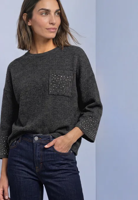 Vrouw draagt donkergrijze trui met glinsterende zak en donkere jeans voor een lichtblauwe achtergrond.