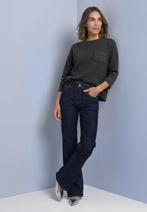 Vrouw leunt tegen muur, grijze trui, donkerblauwe wijde jeans en zilveren schoenen.