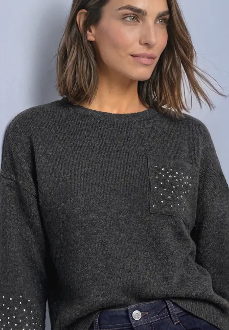 Vrouw in grijze gebreide trui met studs op de zak en jeans, kijkt opzij.