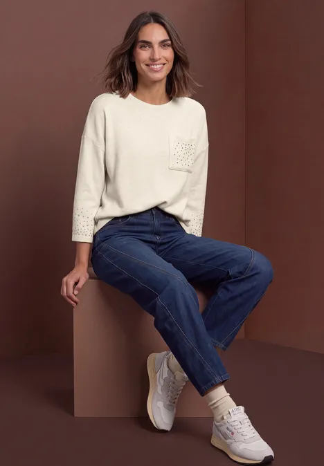 Shirt met 3/4-mouwen en siersteentjes clay sand mel. Eine Frau in lässiger Kleidung: helle 3/4-Arm-Bluse, dunkle Jeans und weiße Sneakers vor braunem Hintergrund.