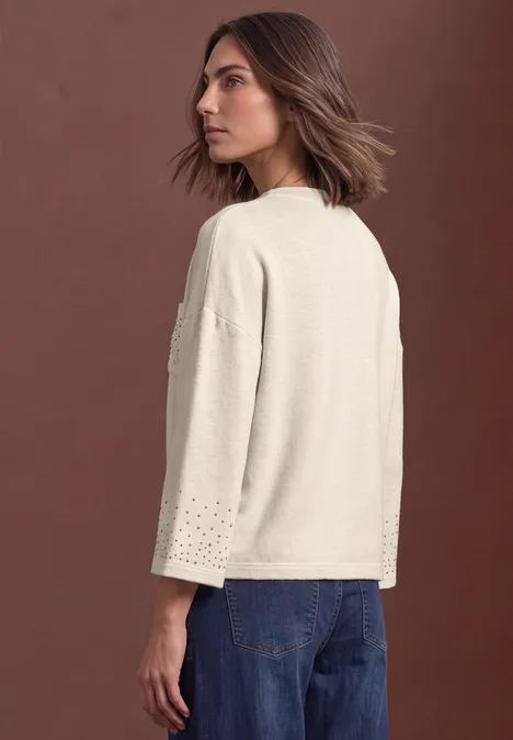Shirt met 3/4-mouwen en siersteentjes clay sand mel. Frau in beigem, lockerem Pullover mit Lochstickerei an den Ärmeln, seitlich stehend vor braunem Hintergrund.