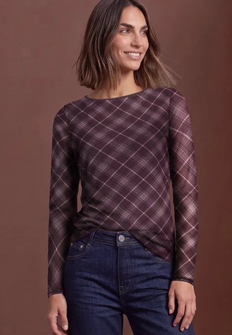 Femme souriante portant un top à carreaux violets à manches longues et un jean, fond uni.