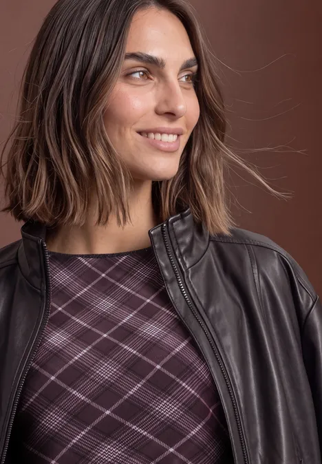 Une femme sourit de profil, portant une veste en cuir marron et un haut à carreaux violets.