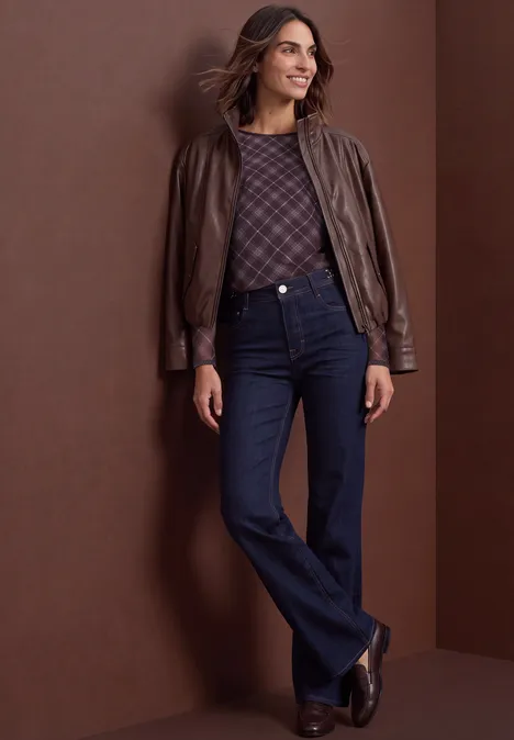 Femme en veste en cuir marron, pull à motif, jean flare foncé et bottines devant un mur brun.