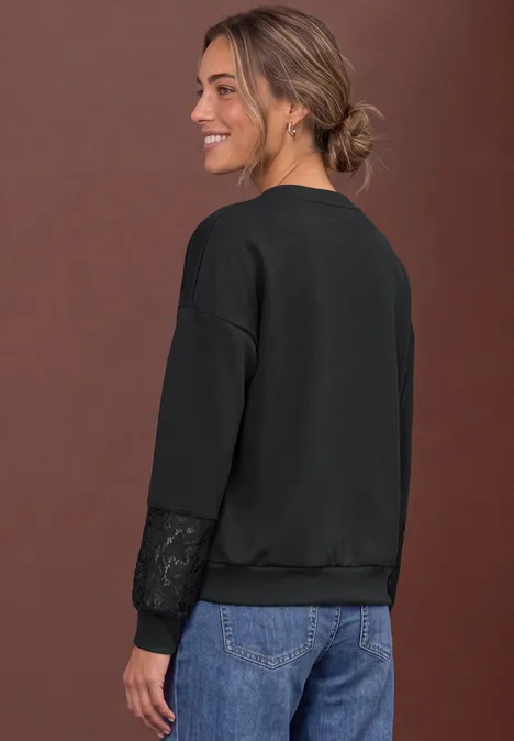 Frau in seitlicher Ansicht trägt schwarzen Pullover mit Spitzen-Details am Ärmel und blaue Jeans, lächelnd.