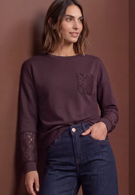 Sweat-shirt à détails dentelle berry plum Frau in lila Langarmshirt mit Spitzenbesatz an Tasche und Ärmeln, dunkle Jeans, vor braunem Hintergrund.