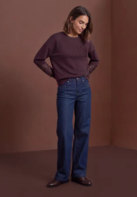 Sweat-shirt à détails dentelle berry plum Frau im lila Pullover mit Spitzenärmeln, weiten Jeans und schwarzen Schuhen vor braunem Hintergrund.