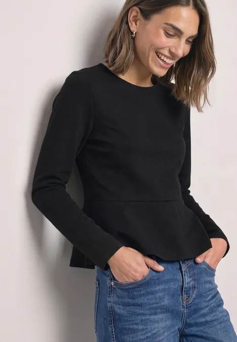 Langarm Shirt mit Volant-Saum Black Eine lächelnde Frau in schwarzem Pullover und Jeans schaut seitlich nach unten.