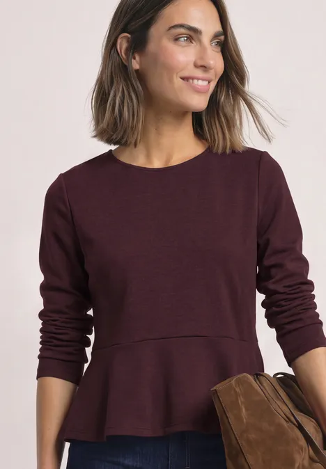 Langarm Shirt mit Volant-Saum berry plum Frau mit brauner Tasche trägt burgunderfarbenes Langarmshirt mit Schößchen-Detail und lächelt.