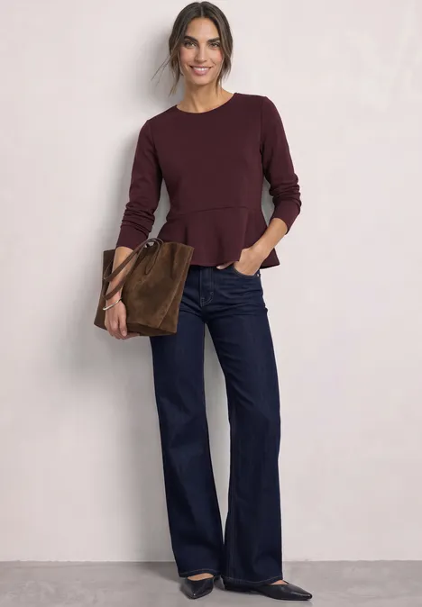 Langarm Shirt mit Volant-Saum berry plum Frau in burgunderrotem Peplum-Oberteil, weiten Jeans und brauner Tasche vor neutralem Hintergrund.
