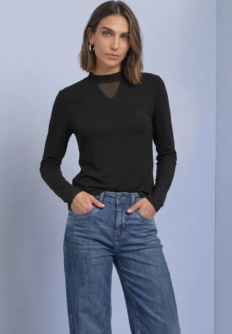 Shirt met lange mouwen, col en mesh-detail Black Frau in schwarzem Langarmshirt mit transparentem Einsatz am Ausschnitt und blauer Jeans, Hände in Taschen.
