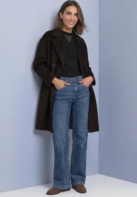 Shirt met lange mouwen, col en mesh-detail Black Frau mit braunem Mantel, schwarzem Shirt, blauen Jeans und braunen Schuhen vor hellem Hintergrund.