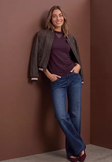 Shirt met lange mouwen, col en mesh-detail berry plum Frau lehnt an brauner Wand, trägt Jeans, violettes Shirt und Jacke über den Schultern, lächelt.