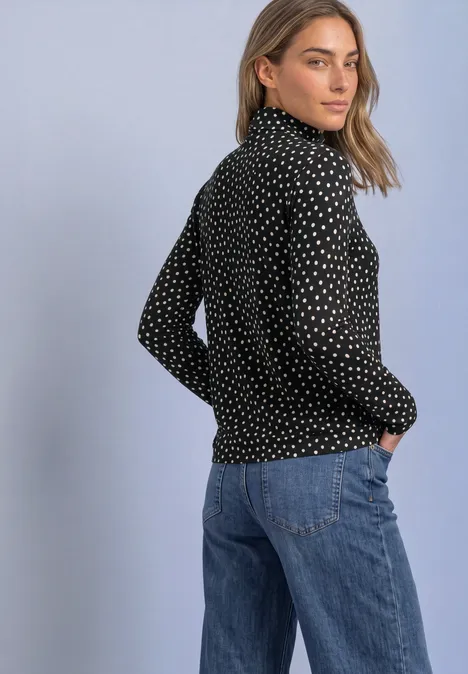 Eenvoudig shirt met col en patroon Black Vrouw in een zwarte coltrui met gouden stippen en een blauwe spijkerbroek, zijwaarts tegen neutrale achtergrond.