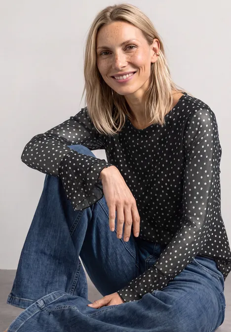 Shirt aus Mesh mit Print und Volantdetails Black Eine lächelnde blonde Frau sitzt locker in Jeans und schwarzer Bluse mit weißen Punkten vor neutralem Hintergrund.