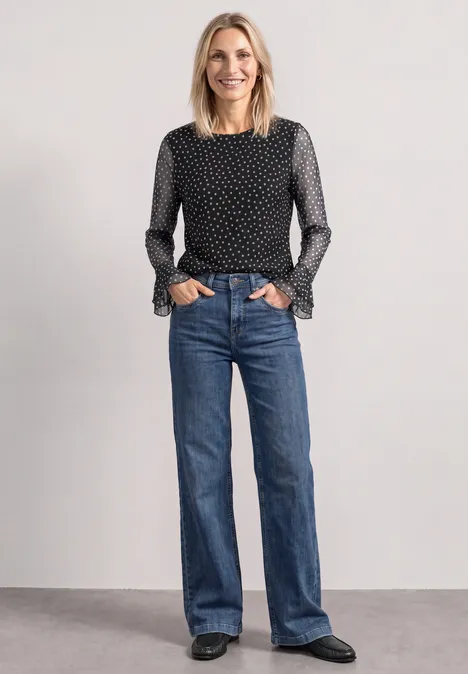Shirt aus Mesh mit Print und Volantdetails Black Frau in schwarz-weiß gepunkteter Bluse mit Rüschenärmeln und weiten blauen Jeans schwarze Schuhe vor grauem Hintergrund.