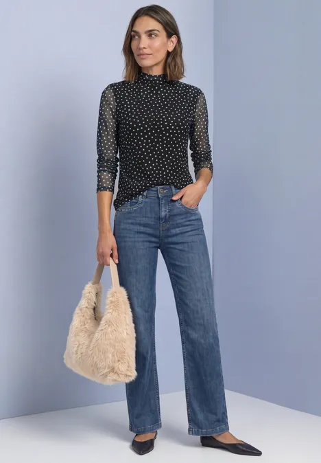 Shirt aus Mesh mit Turtle Neck und Print Black Frau mit schwarzer, gepunkteter Bluse, blauen Jeans und flauschiger Tasche vor schlichtem Hintergrund.