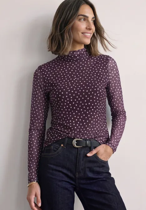 Frau mit dunkelviolettem, gepunktetem Rollkragenpullover und Jeans, lächelnd mit Händen in den Taschen.
