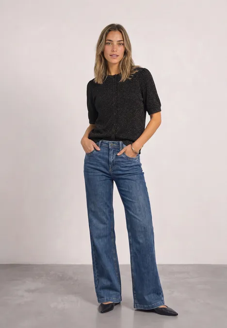 Kurzarm Jacke mit Rundhals und Glitzer Black Frau in schwarzem Pullover und weiten blauen Jeans steht vor neutralem Hintergrund, Hände in den Taschen.