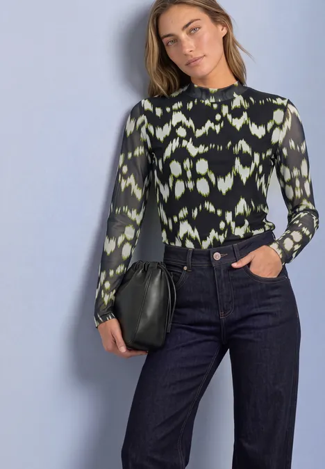 Shirt van mesh met col en print Black Frau trägt schwarz-grün gemustertes Langarmshirt, weite Jeans und hält eine schwarze Clutch vor hellem Hintergrund