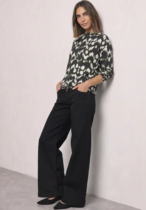 Shirt mit Turtle Neck und Tunnelzug Black Frau mit weiten schwarzen Hosen und abstrakt gemustertem Oberteil steht lässig vor hellem Hintergrund.