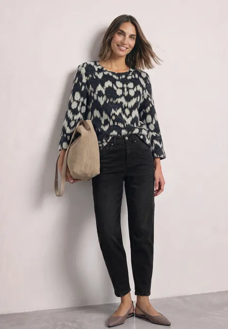 Une femme souriante porte un jean taille haute noir, un top à motifs et tient un sac beige.