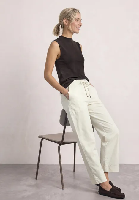 Basic top espresso brown Frau in schwarzem Top und weißen Hosen lehnt lässig an einem Stuhl.