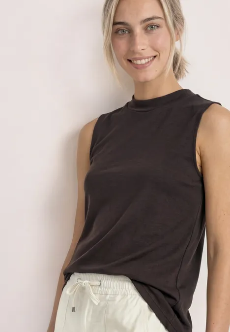 Basic top espresso brown Eine lächelnde Frau in schwarzem Ärmellosen Top und weißen Hose vor neutralem Hintergrund.