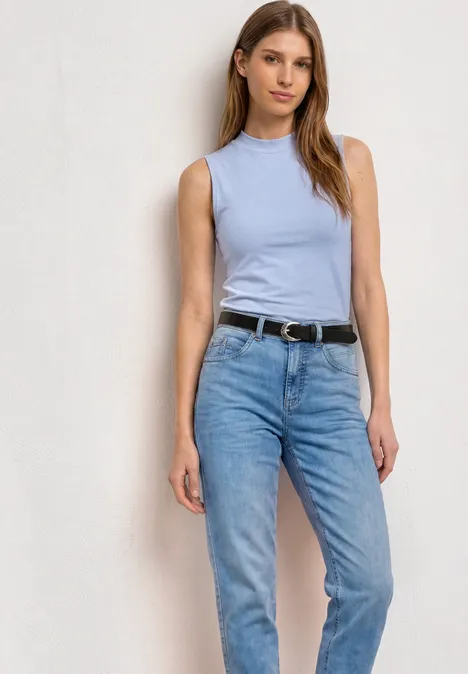 Frau in blauem Top und Jeans mit Gürtel vor hellem Hintergrund.