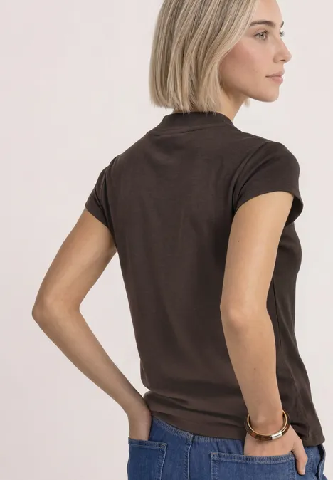 Frau im Profil mit blondem Bob trägt braunes T-Shirt und Jeans vor neutralem Hintergrund.