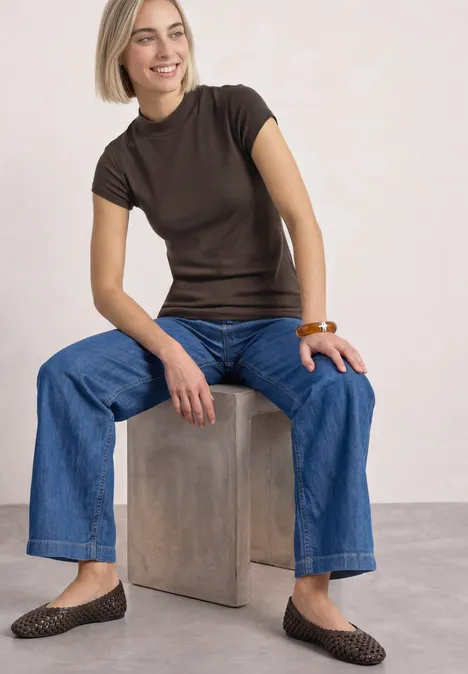 Frau mit blondem Bob sitzt auf einem Block, trägt braunes T-Shirt, weite Jeans, braune geflochtene Ballerinas und Holzarmreif.