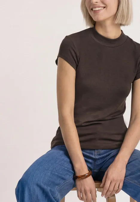 Frau sitzt lächelnd, trägt braunes T-Shirt, blaue Jeans und Holzarmband.