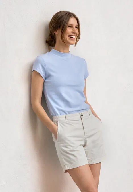 Basic Shirt mit Turtle Neck cashmere blue Eine junge Frau lehnt an einer Wand, trägt hellblauen Kurzarmpulli und beige Shorts und lächelt.