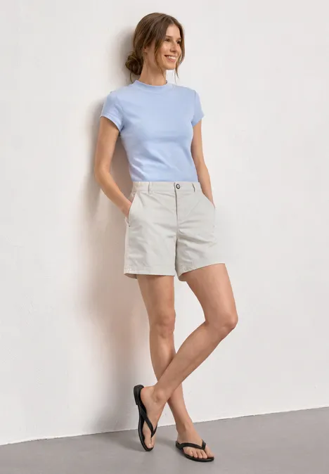 Basic Shirt mit Turtle Neck cashmere blue Frau in hellblauem T-Shirt, beigen Shorts und schwarzen Flip-Flops lehnt lässig an weißer Wand.