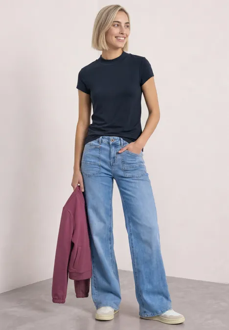 Basic Shirt mit Turtle Neck shadowed navy Frau trägt schwarzes T-Shirt, weite Jeans und weiße Sneakers, hält eine rosa Jacke.