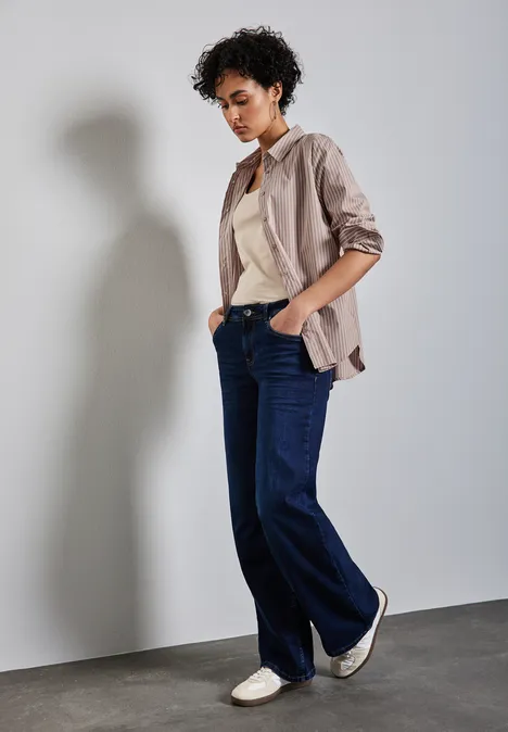 Frau in weiter Jeans, gestreiftem Hemd und Sneakers steht lässig an der Wand.
