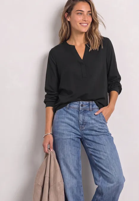 Frau mit schwarzer Bluse und weiter blauer Jeans hält eine Jacke und lächelt.