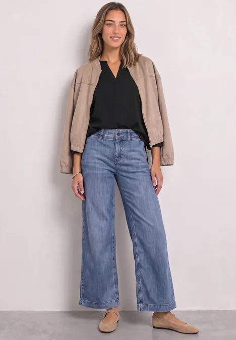 Frau in lockerer Jeans mit weitem Bein, schwarzem Top und beiger Jacke, lässig mit hellen Slippern.