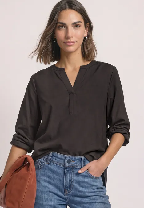 Frau in schwarzer Bluse mit V-Ausschnitt, hochgekrempelten Ärmeln und Jeans, die eine braune Handtasche hält.
