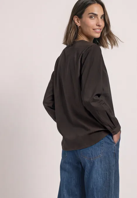 Frau in dunkler Bluse und blauen Jeans dreht sich leicht zur Kamera, Hände in den Taschen.