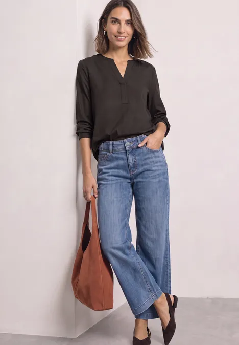 Frau in schwarzem Langarmshirt, blauen Jeans und brauner Tasche lehnt an weißer Wand.