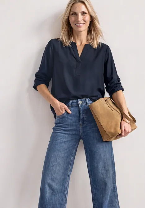 Split Neck Tunika darkest blue Frau in dunkelblauer Bluse und weitem Jeans, mit brauner Tasche, steht lächelnd vor heller Wand.