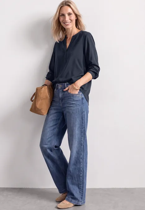 Split Neck Tunika darkest blue Frau in dunkler Bluse und weitem Jeans vor hellem Hintergrund mit brauner Tasche.