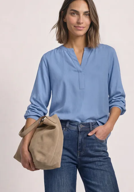 Frau in hellblauer Bluse und Jeans hält eine beige Tasche, vor schlichter Kulisse.