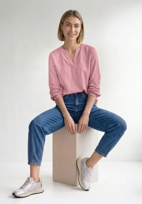 Eine Frau sitzt lächelnd auf einem Würfel, trägt pinke Bluse, blaue Jeans und weiße Sneaker vor weißem Hintergrund.
