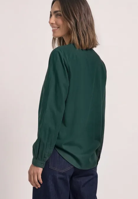 Frau in dunkelgrüner Bluse und Jeans, seitlich stehend und lächelnd.