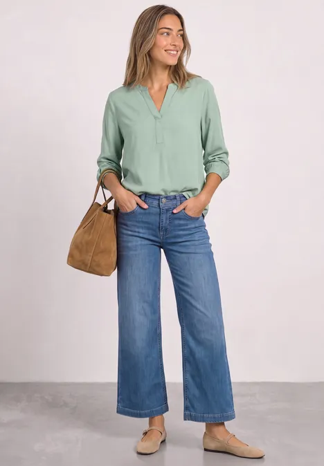 Lachende vrouw in groen blouse, wijdvallende jeans, lichte schoenen en bruine tas voor neutrale achtergrond.