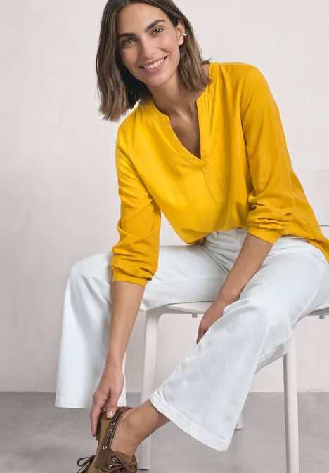 Vrouw in gele blouse en witte broek zit ontspannen en strikt haar veters.